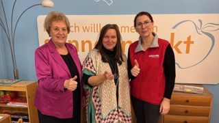 Drei Frauen stehen lächelnd vor einem Banner. Die Frau links trägt einen rosa Blazer, die mittlere ein gemustertes Tuch und die rechte eine rote Weste mit der Aufschrift 'volkshilfe'.