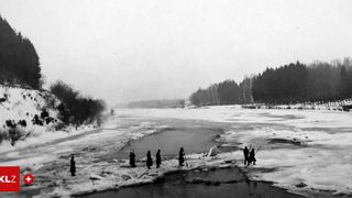 Schwarz-Weiß-Bild von Menschen, die auf schneebedecktem Eis stehen. Mehrere Bäume säumen die Flussufer.
