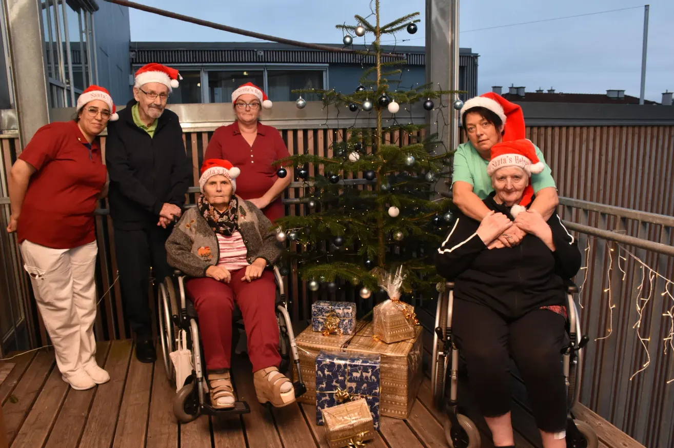 Eine Gruppe von Menschen, darunter ältere Personen in Rollstühlen, posiert für ein Foto vor einem geschmückten Weihnachtsbaum mit Geschenken auf einer Holzterrasse.
