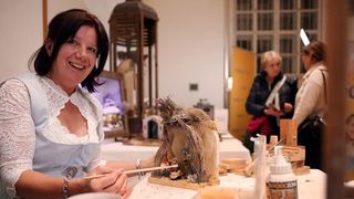 Eine Frau malt eine Skulptur eines Paares von Tieren, lächelt in einem Kunstatelier mit einer Holzlampe und einem Fenster.