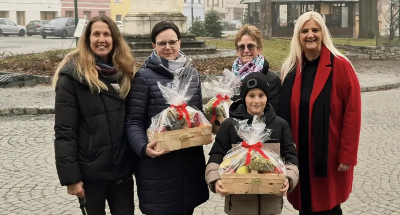 Fünf Menschen stehen in einem Außenbereich und lächeln. Sie halten Obstkörbe mit roten Bändern in der Hand. Im Hintergrund ist ein verschwommener Hintergrund mit einer Statue, Gebäuden und einem Baum zu sehen.