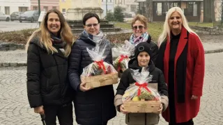 Fünf Menschen stehen in einem Außenbereich und lächeln. Sie halten Obstkörbe mit roten Bändern in der Hand. Im Hintergrund ist ein verschwommener Hintergrund mit einer Statue, Gebäuden und einem Baum zu sehen.