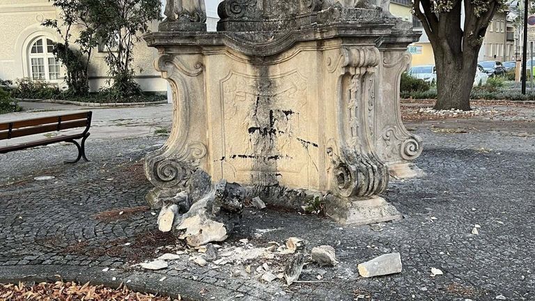 Eine alte Steinstruktur mit kunstvollen Schnitzereien und einem beschädigten Schild, umgeben von Schutt, mit einem Baum und einem Gebäude im Hintergrund.