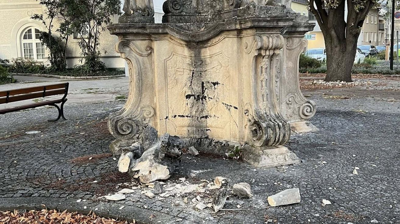 Eine alte Steinstruktur mit kunstvollen Schnitzereien und einem beschädigten Schild, umgeben von Schutt, mit einem Baum und einem Gebäude im Hintergrund.
