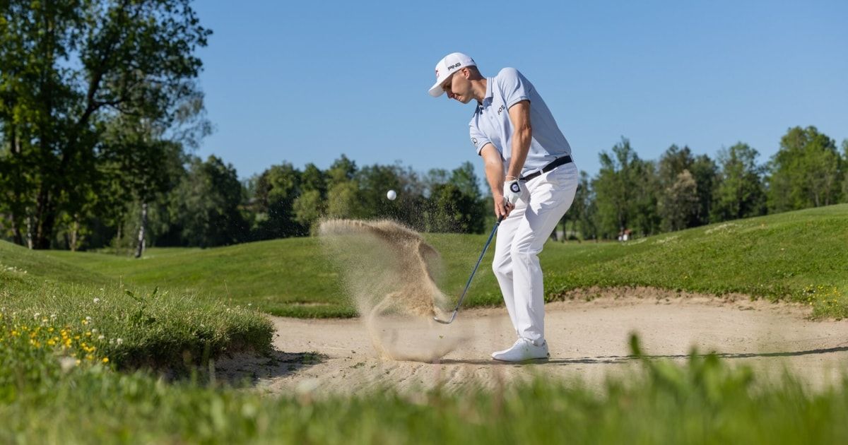 Ein Golfer in einem blauen Shirt und einer weißen Mütze schlägt einen Golfball aus einem sandigen Bunker auf einem Golfplatz.