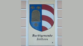 Ein Schild mit blau-weiß gestreiftem Hintergrund, einem Bienenstock-Design und zwei Bienen, zeigt den Text 'Marktgemeinde Zellern' in schwarzer Schrift.