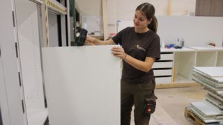 Eine Frau in einem braunen T-Shirt hält einen Hammer und arbeitet in einer Werkstatt an einer weißen Tafel.