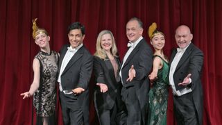 Fünf Menschen in formeller Kleidung posieren für ein Foto auf einer Bühne mit einem roten Vorhang im Hintergrund.