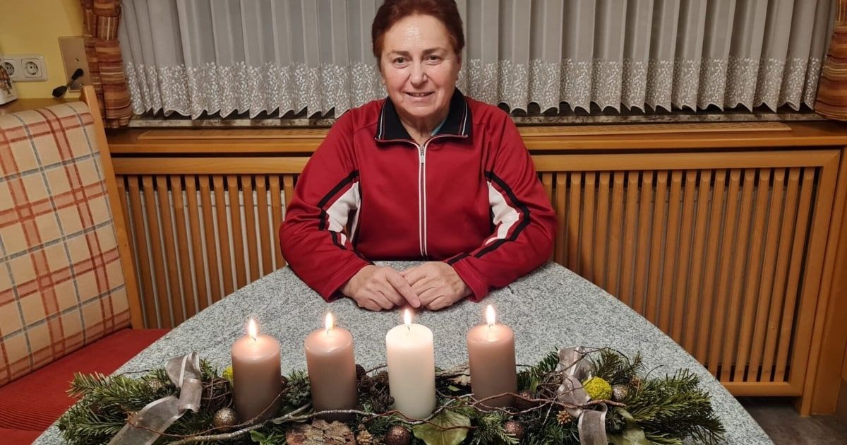 Eine Frau sitzt an einem Tisch mit vier brennenden Kerzen vor ihr. Sie trägt eine rote Jacke und lächelt in die Kamera. Hinter ihr ist ein weißer Vorhang und eine Holzwand.