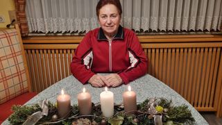 Eine Frau sitzt an einem Tisch mit vier brennenden Kerzen vor ihr. Sie trägt eine rote Jacke und lächelt in die Kamera. Hinter ihr ist ein weißer Vorhang und eine Holzwand.