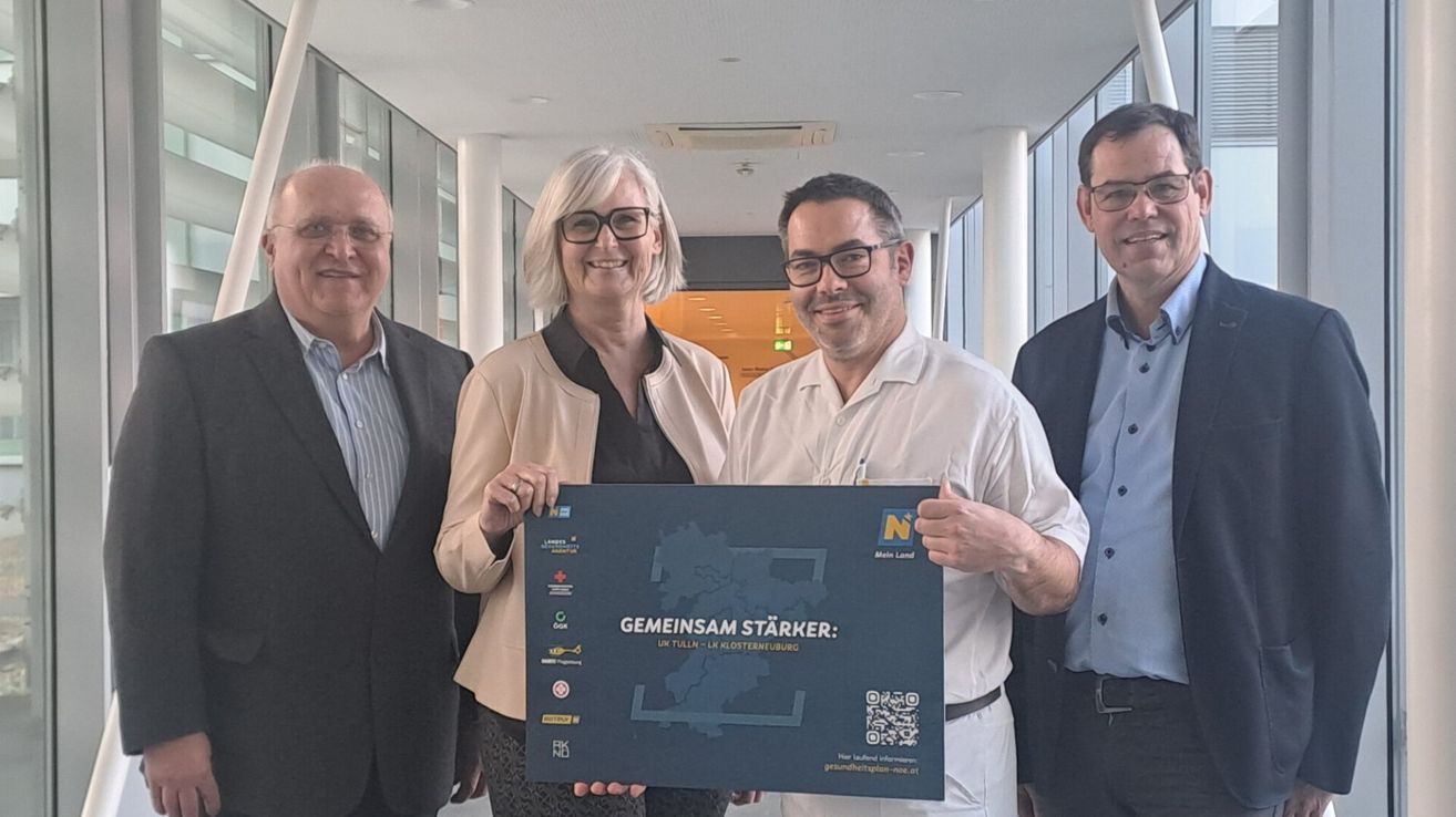 Vier Personen stehen in einem Korridor und lächeln. Eine Frau und ein Mann in Weiß halten ein blaues Schild mit einer Karte und Text, während ein anderer Mann mit Brille neben ihnen lächelt.