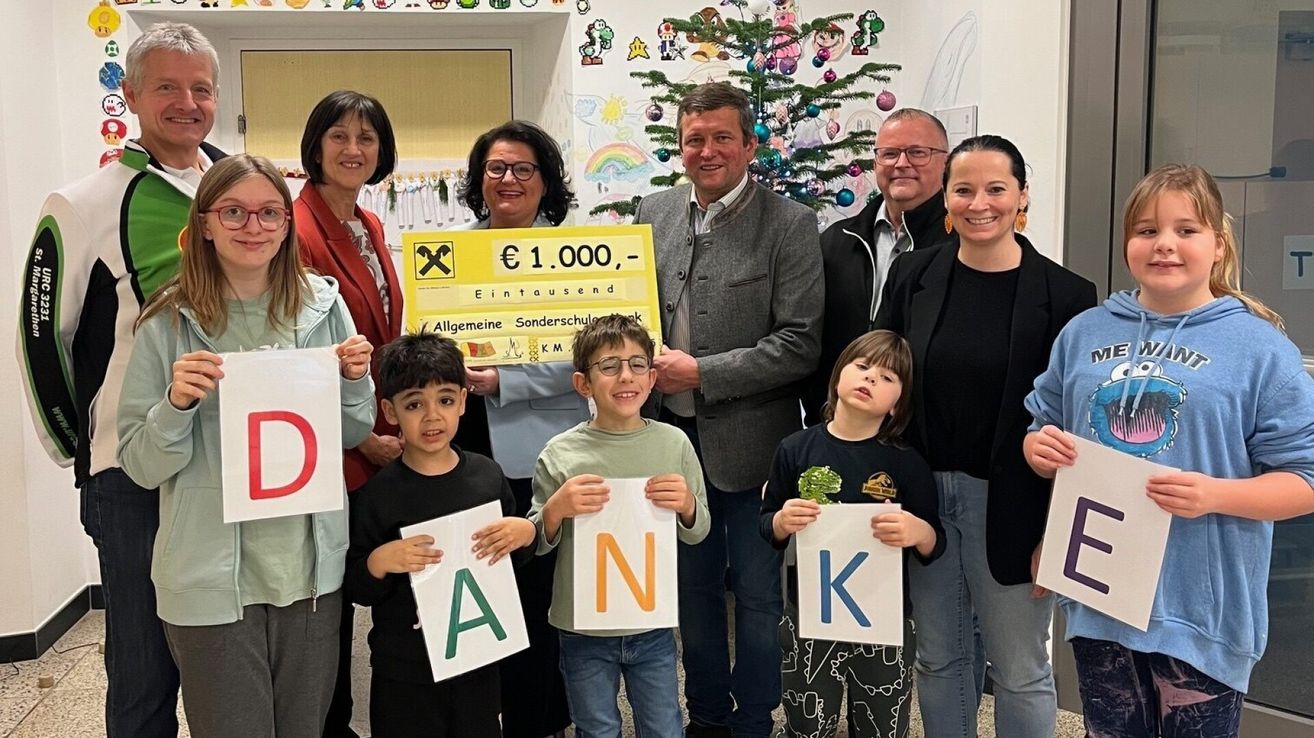 Mehrere Menschen stehen in einem Raum, wahrscheinlich einem Klassenzimmer, und halten Schilder mit Buchstaben und einem Scheck. Sie lächeln und posieren für ein Foto.