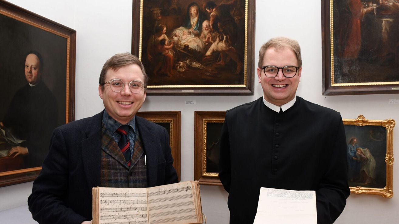 Zwei Männer stehen in einem Raum mit Kunstwerken, einer hält ein Musikbuch und der andere ein Dokument. Sie lächeln und posieren für ein Foto.