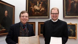 Zwei Männer stehen in einem Raum mit Kunstwerken, einer hält ein Musikbuch und der andere ein Dokument. Sie lächeln und posieren für ein Foto.