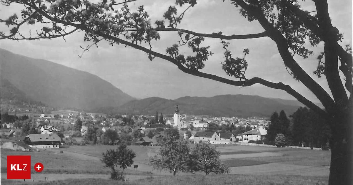 Ein Schwarz-Weiß-Foto einer kleinen Stadt in einem Tal mit Bergen im Hintergrund. Die Stadt hat einen Kirchturm und ist von Bäumen und Feldern umgeben.