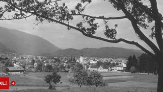 Ein Schwarz-Weiß-Foto einer kleinen Stadt in einem Tal mit Bergen im Hintergrund. Die Stadt hat einen Kirchturm und ist von Bäumen und Feldern umgeben.
