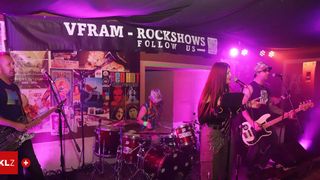 Eine Band tritt auf der Bühne unter einem Banner mit der Aufschrift 'Vfram - Rockshows Follow Us' bei lila Beleuchtung auf. Eine Frau singt in ein Mikrofon, während zwei Männer Gitarre und Schlagzeug spielen.