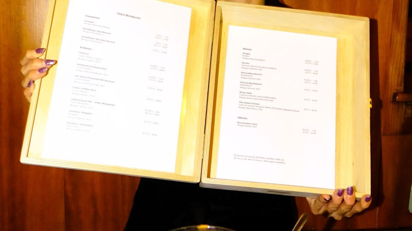 Geöffnete Holzbox mit einem Weinmenü, wobei zwei Seiten sichtbar sind. Die erste Seite listet verschiedene Weinnamen und Preise auf. Die zweite Seite hat den Titel 'Valo Weinkarte' und beinhaltet Weinbeschreibungen und Preise.