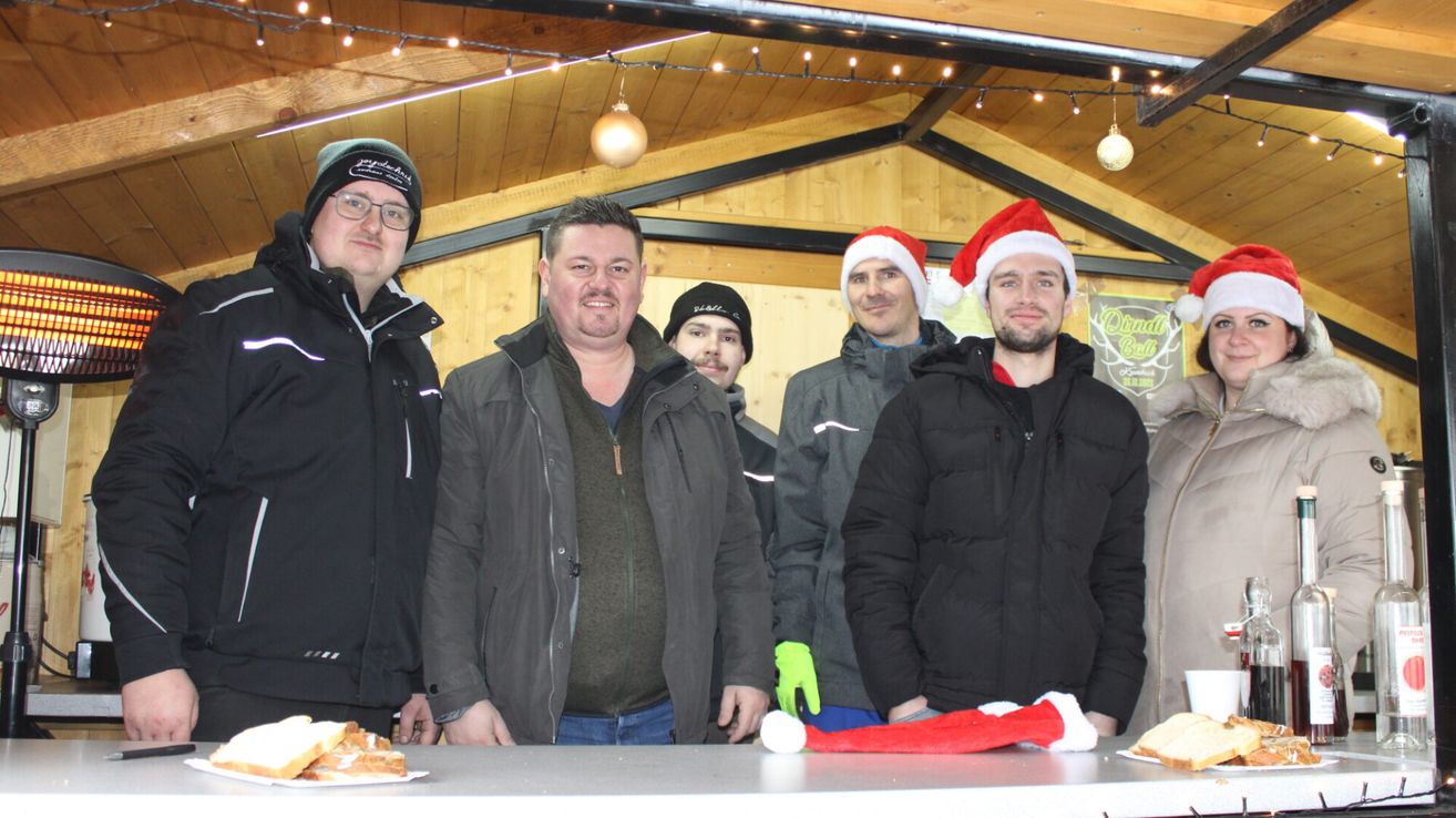 Fünf Männer stehen in Winterkleidung und Weihnachtsmützen im Innenraum. Sie lächeln für die Kamera, wobei Essen und ein Weihnachtsbaum im Hintergrund zu sehen sind.
