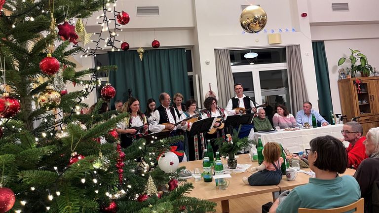 Eine festliche Zusammenkunft von Menschen, die Musik in der Nähe eines geschmückten Weihnachtsbaums genießen, mit Musikern, die Saiteninstrumente spielen. Ein Tisch mit Getränken und Essen ist aufgebaut, und eine Frau sitzt auf einem Stuhl.