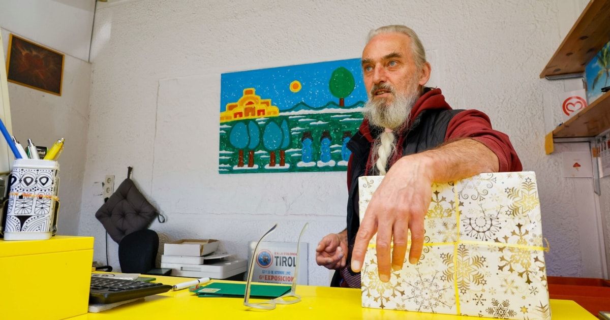 Ein Mann mit weißem Bart hält ein verpacktes Geschenk in einem Raum mit einem Schreibtisch.