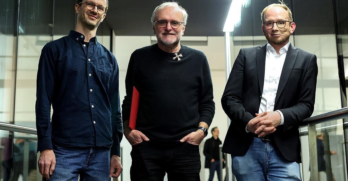 Drei Männer stehen zusammen in einem Raum. Der Mann auf der linken Seite trägt ein blaues Hemd und Jeans. Der Mann in der Mitte trägt einen schwarzen Pullover und hält eine rote Mappe. Der Mann auf der rechten Seite trägt einen Anzug. Alle drei Männer tragen eine Brille.