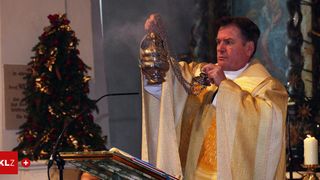 Ein Priester hält eine goldene Lampe und scheint an einem Podium zu sprechen, mit einem Weihnachtsbaum dahinter.
