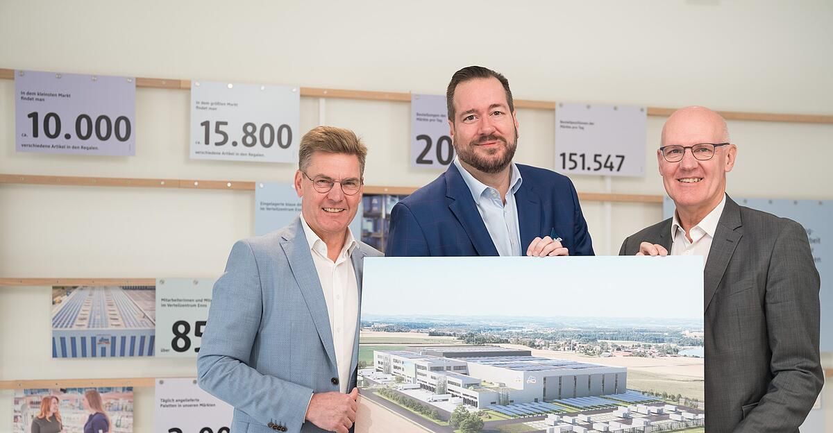 Drei Männer stehen vor einem Brett mit Zahlen und einem Gebäudemodell, lächeln und posieren für ein Foto.