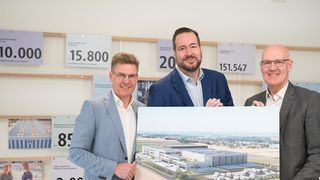 Drei Männer stehen vor einem Brett mit Zahlen und einem Gebäudemodell, lächeln und posieren für ein Foto.