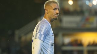 Ein männlicher Fußballspieler mit kurzem Haarschnitt steht auf dem Spielfeld und schaut nach links. Er trägt ein weißes Trikot mit blauen Streifen.