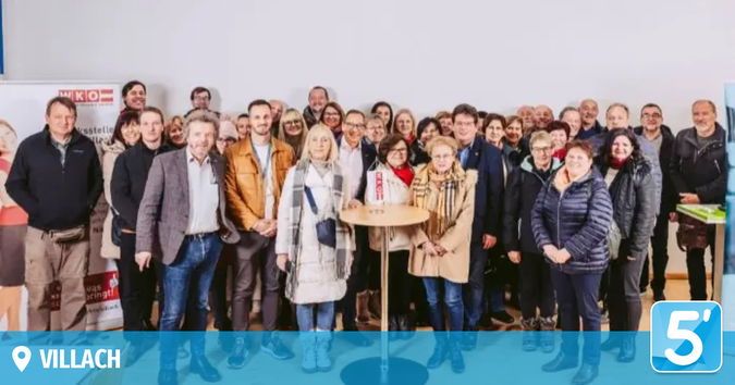 Eine Gruppe Erwachsener in Winterkleidung, die für ein Foto in einem Raum mit weißer Wand, kleinem Tisch und einem Schild im Hintergrund posiert.