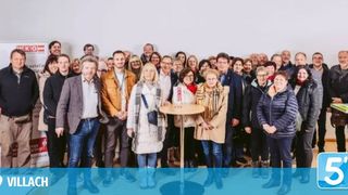 Eine Gruppe Erwachsener in Winterkleidung, die für ein Foto in einem Raum mit weißer Wand, kleinem Tisch und einem Schild im Hintergrund posiert.