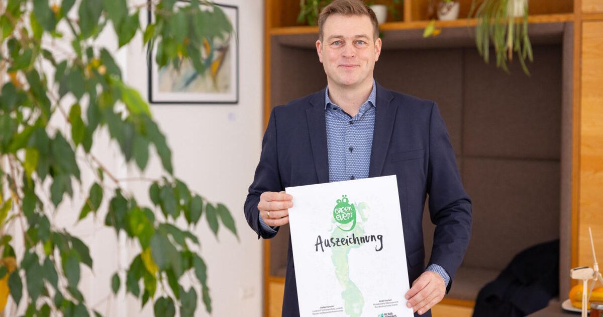 Ein Mann in einem Anzug hält eine Urkunde mit einem grünen Fußabdruck und dem Wort 'Auszeichnung'. Dahinter eine grüne Pflanze und eine Wand mit einem Bilderrahmen.