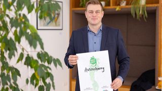 Ein Mann in einem Anzug hält eine Urkunde mit einem grünen Fußabdruck und dem Wort 'Auszeichnung'. Dahinter eine grüne Pflanze und eine Wand mit einem Bilderrahmen.