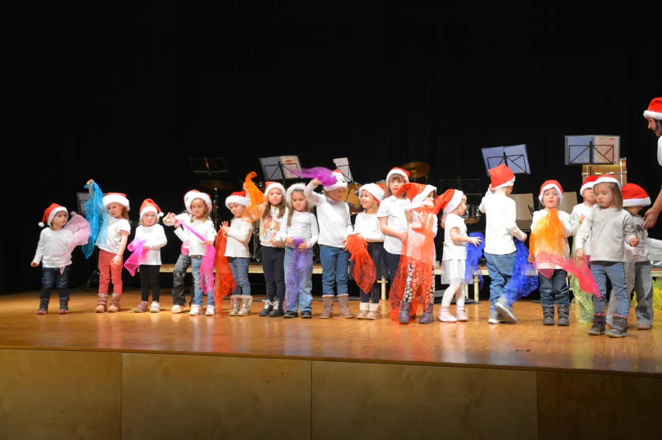 Eine Gruppe von Kindern mit Weihnachtsmützen tritt auf der Bühne auf. Einige halten bunte Requisiten. Hinter ihnen sind Musikinstrumente aufgestellt.