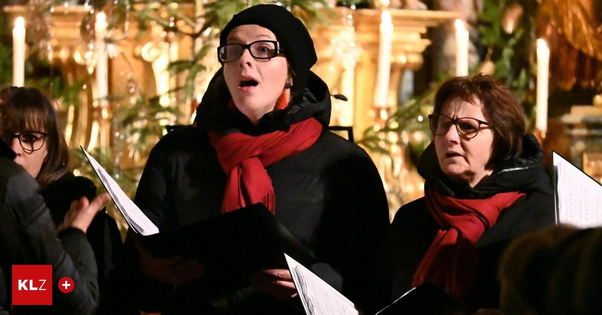 Zwei Frauen, beide mit roten Schals, stehen und singen in einer Kirche mit Weihnachtsdekoration.