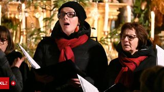 Zwei Frauen, beide mit roten Schals, stehen und singen in einer Kirche mit Weihnachtsdekoration.