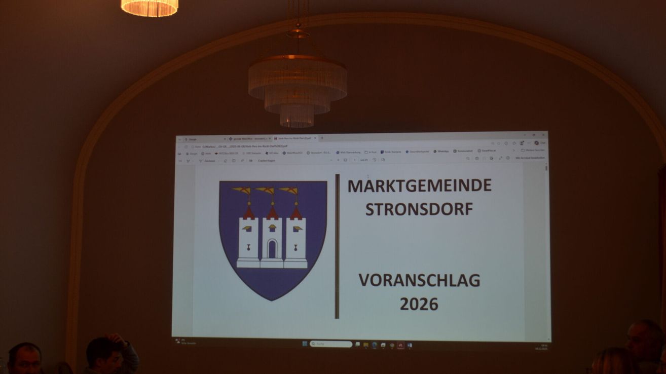 Eine Präsentationsfolie zeigt einen Wappenschild mit dem Text 'MARKTGEMEINDE STRONSDORSF VORANSCHLAGE 2026' auf einem Bildschirm, mit einer Kronleuchterlampe, die von der Decke hängt.