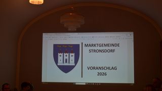 Eine Präsentationsfolie zeigt einen Wappenschild mit dem Text 'MARKTGEMEINDE STRONSDORSF VORANSCHLAGE 2026' auf einem Bildschirm, mit einer Kronleuchterlampe, die von der Decke hängt.