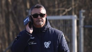 Ein Mann mit Sonnenbrille und einer dunkelblauen Jacke mit weißem Logo steht draußen und hält ein schwarzes Gerät in der linken Hand.