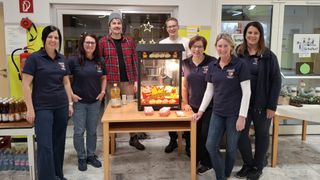 Eine Gruppe von Menschen steht um eine Popcornmaschine und lächelt. Ein Tisch hält eine Flasche und Popcorn-Kisten. Sie tragen Uniformen. Hinter ihnen ist ein Fenster.
