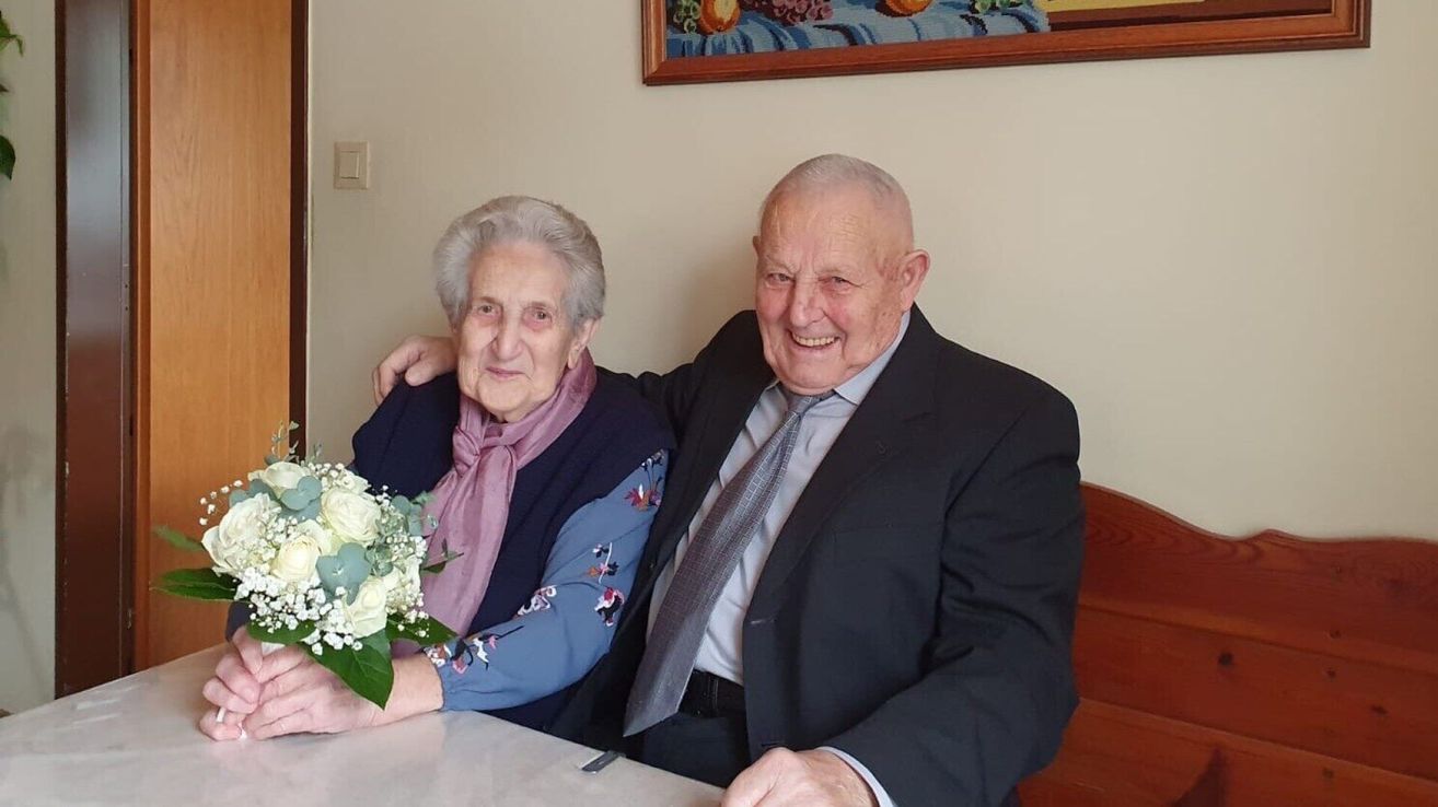 Ein älteres Paar lächelt und posiert für ein Foto, die Frau hält einen Blumenstrauß, der Mann trägt einen Anzug und eine Krawatte, sie sitzen zusammen auf einer Bank.