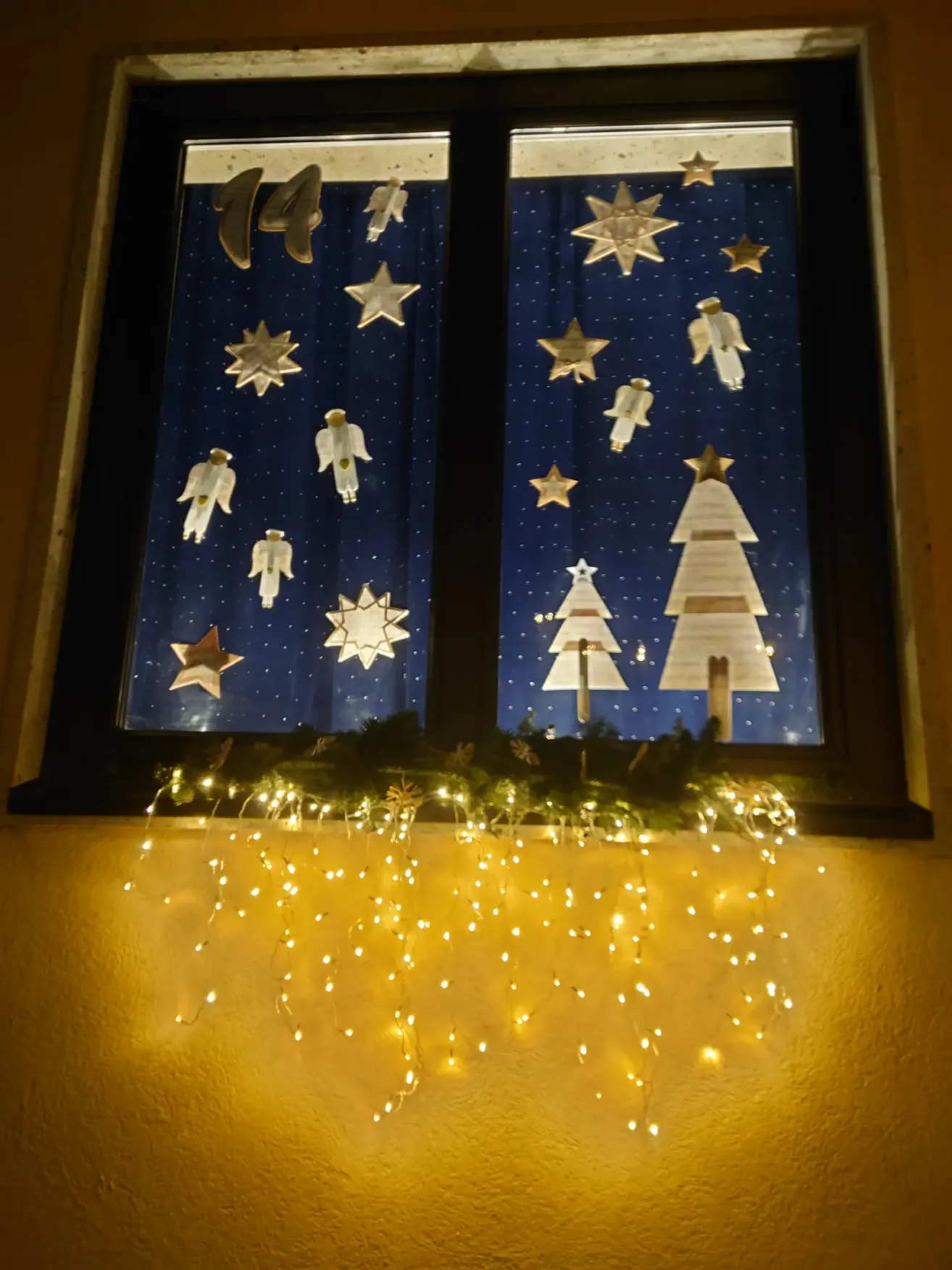 Ein Fenster zeigt eine Weihnachtsdekoration mit Sternen, Engeln und Weihnachtsbäumen. Unten am Fenster befindet sich auch eine Lichterkette.