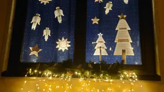 Ein Fenster zeigt eine Weihnachtsdekoration mit Sternen, Engeln und Weihnachtsbäumen. Unten am Fenster befindet sich auch eine Lichterkette.