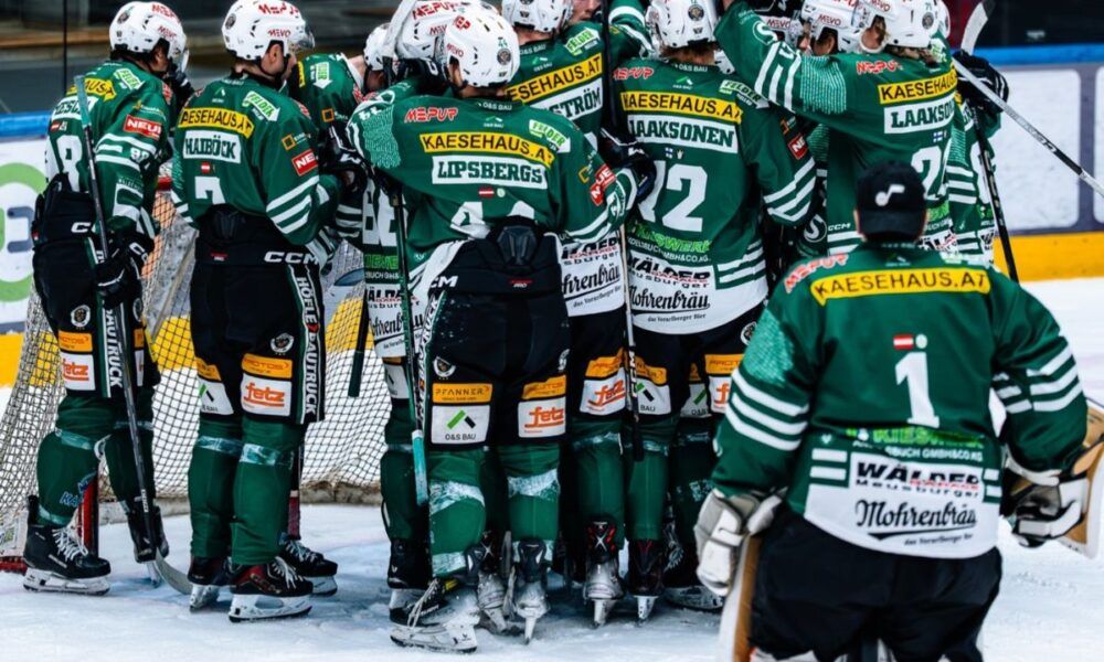 Eine Eishockeymannschaft in grünen Uniformen versammelt sich neben dem Tornetz, mit sichtbaren Nummern und Sponsorenlogos, in Vorbereitung auf ein Spiel.