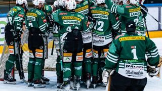 Eine Eishockeymannschaft in grünen Uniformen versammelt sich neben dem Tornetz, mit sichtbaren Nummern und Sponsorenlogos, in Vorbereitung auf ein Spiel.