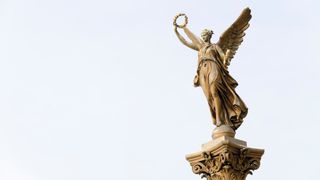 Eine bronzene Engelsstatue, die einen Lorbeerkranz hält, steht auf einer Säule, vor einem grauen Himmel.