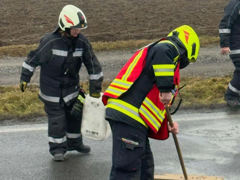 Zwei Feuerwehrleute in voller Ausrüstung gehen auf einer nassen Straße. Einer hält einen weißen Behälter, der andere benutzt einen Stock.