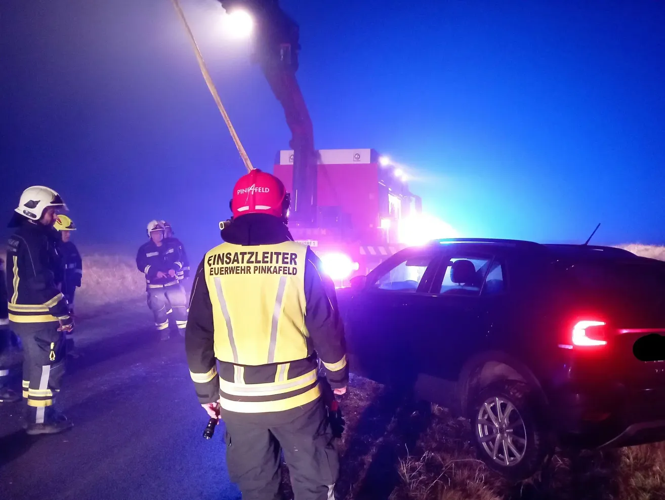 Feuerwehrleute stehen an der Straße bei einem Autounfall. Ein Feuerwehrmann steht mit einer Taschenlampe und trägt eine gelbe Weste. Ein anderer Feuerwehrmann steht hinter einem Auto mit eingeschalteten Warnleuchten.