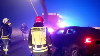 Feuerwehrleute stehen an der Straße bei einem Autounfall. Ein Feuerwehrmann steht mit einer Taschenlampe und trägt eine gelbe Weste. Ein anderer Feuerwehrmann steht hinter einem Auto mit eingeschalteten Warnleuchten.